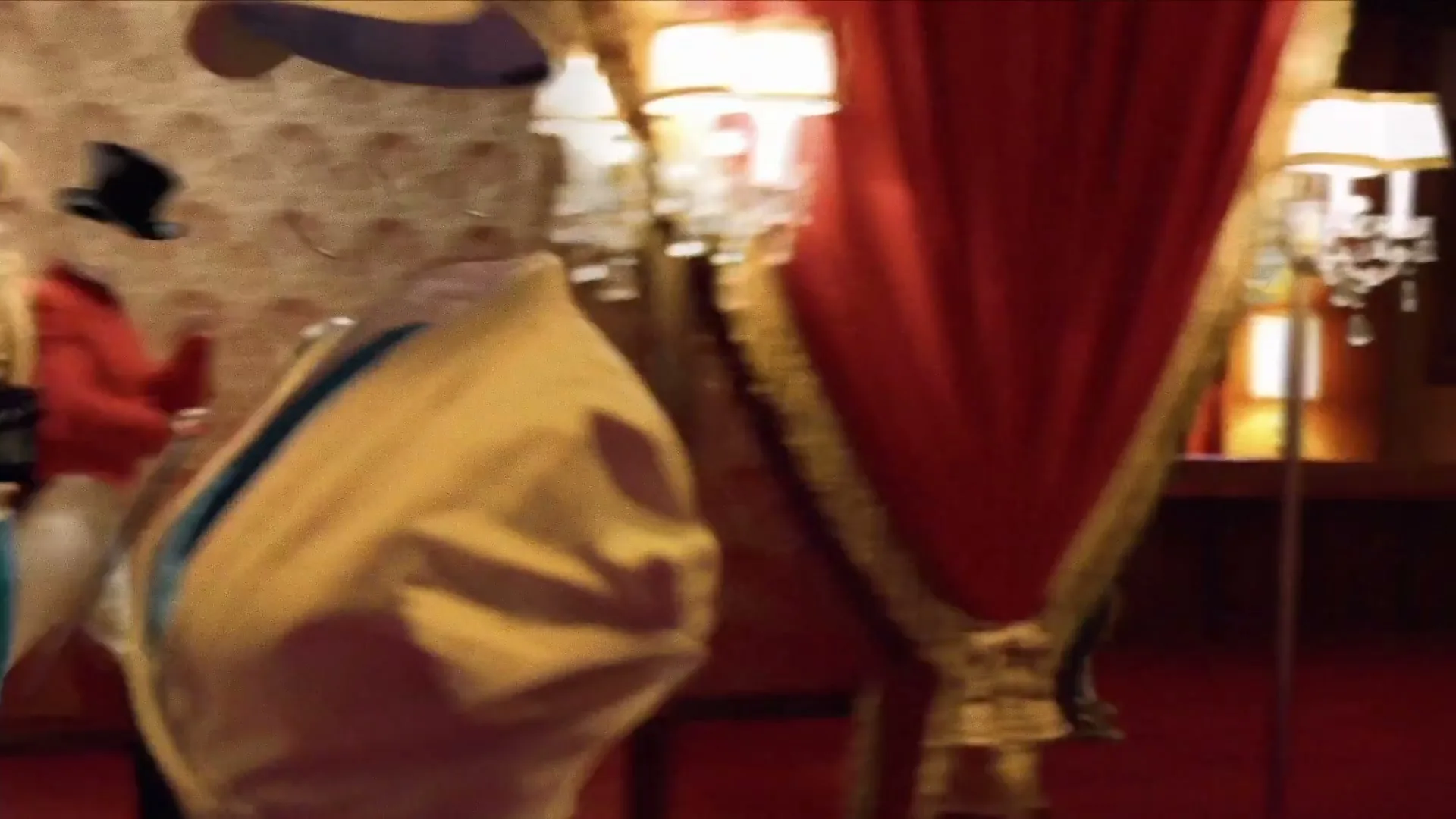 An invisible man, dressed in a red tailcoat, white breeches, black boots, and a top hat, walks with a cane across a red carpet in a luxurious room.