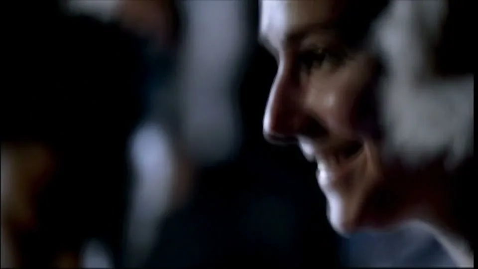 A woman with dark hair smiles, her face illuminated by a soft light, in a dark, indistinct interior setting.