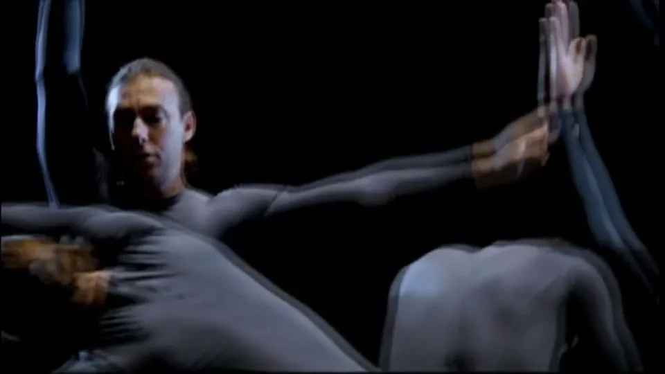 A group of dancers in grey bodysuits perform synchronized movements on a dark stage, creating a dynamic and fluid visual.