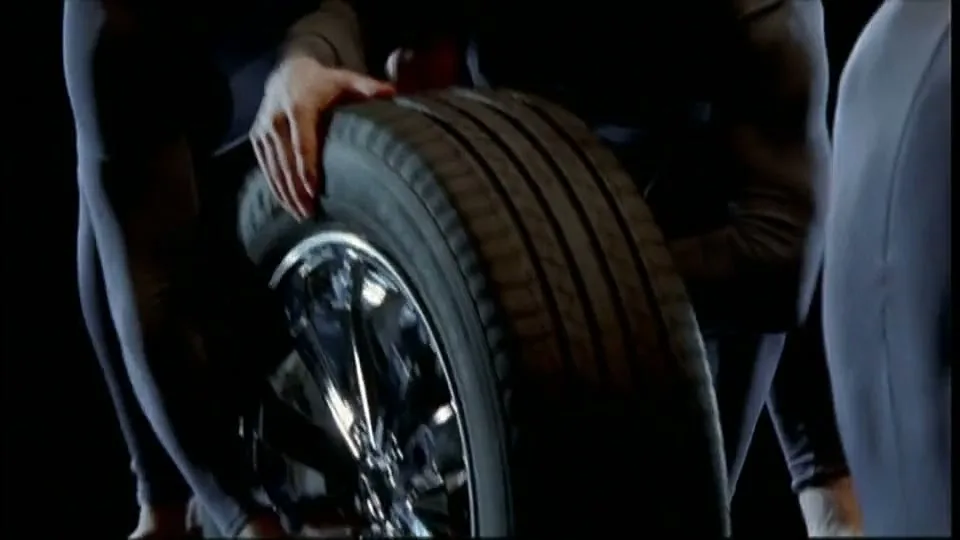 Several people in dark bodysuits assemble a car wheel, with hands and feet visible against a dark background.