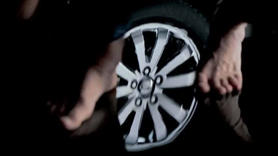 A group of people in grey bodysuits are seen in a kaleidoscope-like reflection, forming a car wheel, in a dark studio.