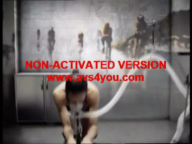 A female athlete in a cycling suit with a breathing mask trains on a stationary bike in a lab, with blurred images of other athletes on the wall.
