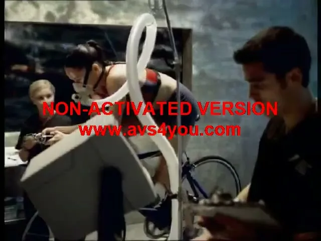 A female athlete in a sports bra and shorts cycles on a stationary bike with a breathing mask, while two scientists monitor her in a lab.