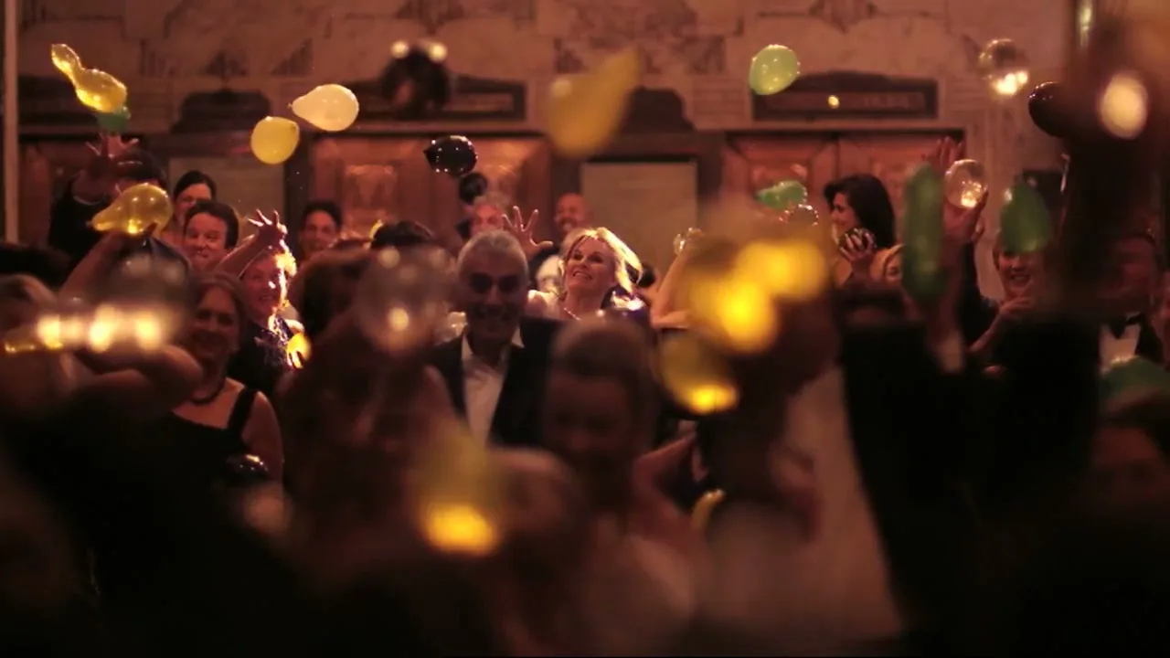A diverse group of adults in formal wear joyfully celebrate as balloons fall around them in a grand, ornate hall.