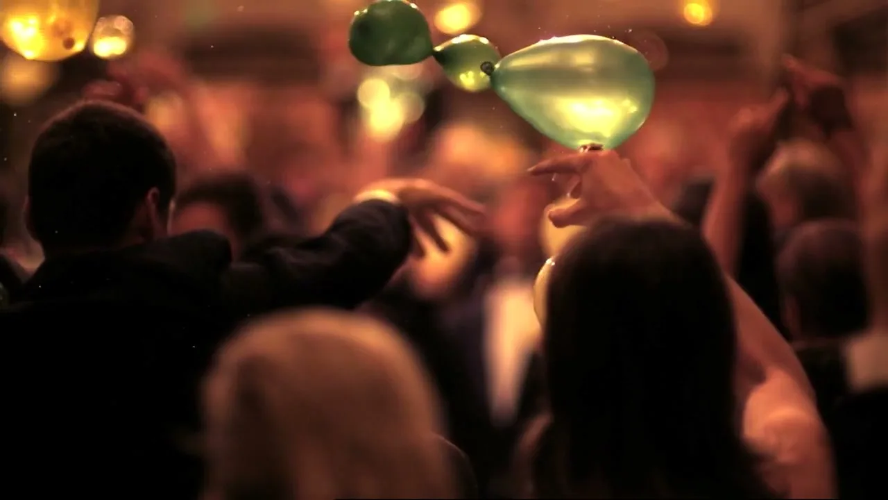 A green balloon floats above a crowd of people dancing at a party, with warm lights in the background.