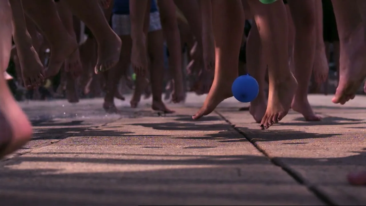 A low-angle shot shows many bare feet dancing on a wet patio, with colorful water balloons bouncing around them in slow motion.