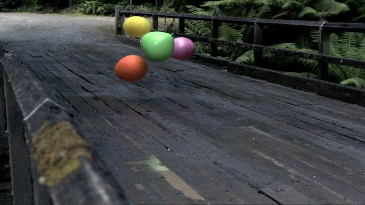 Four colorful, glossy, fruit-shaped objects float above a weathered wooden bridge in a lush, green forest setting.