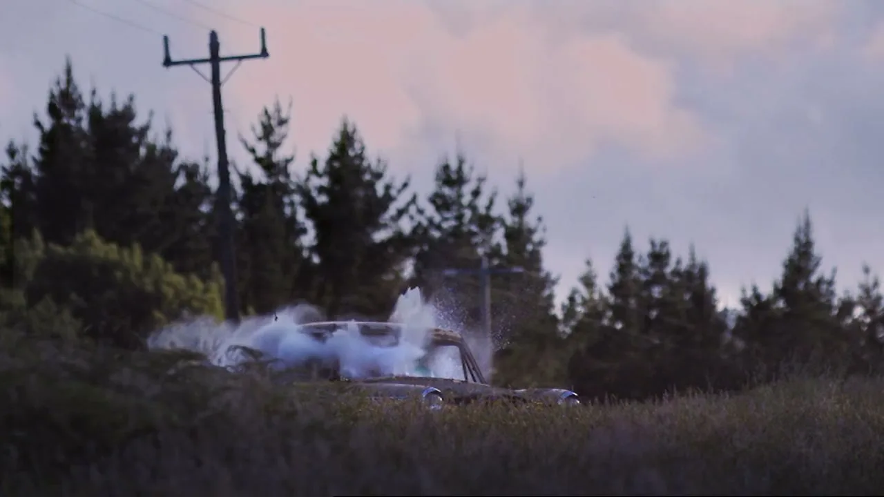 A vintage car, partially submerged in tall grass, emits a burst of water and smoke from its interior, with a forest and power lines in the background under a cloudy sky.
