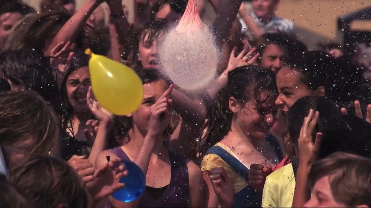 A crowd of diverse teens and children are soaked by water as a pink water balloon bursts above them, with yellow and blue balloons also visible.