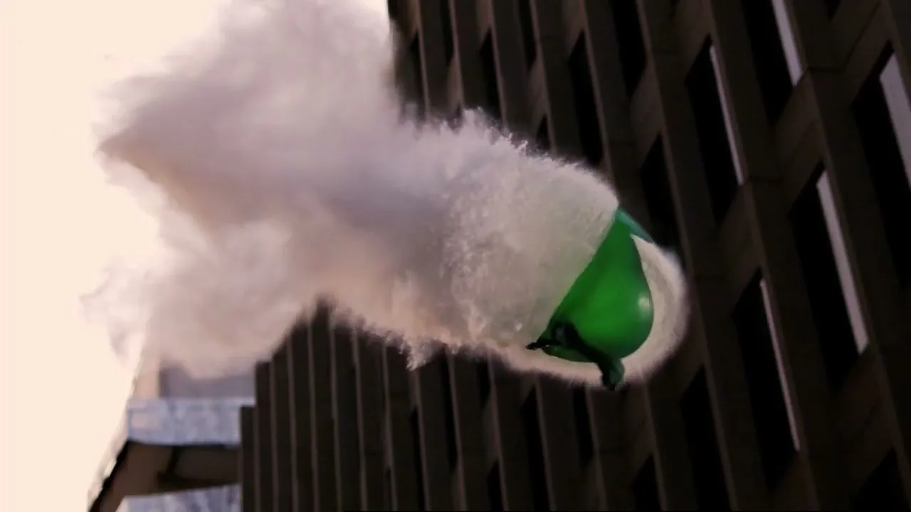 A green balloon bursts in slow motion, releasing a cloud of white powder against the backdrop of a modern building.
