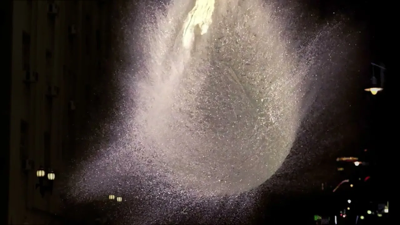 A large water balloon bursts in slow motion, creating a massive splash of water droplets against a dark, blurry city background at night.