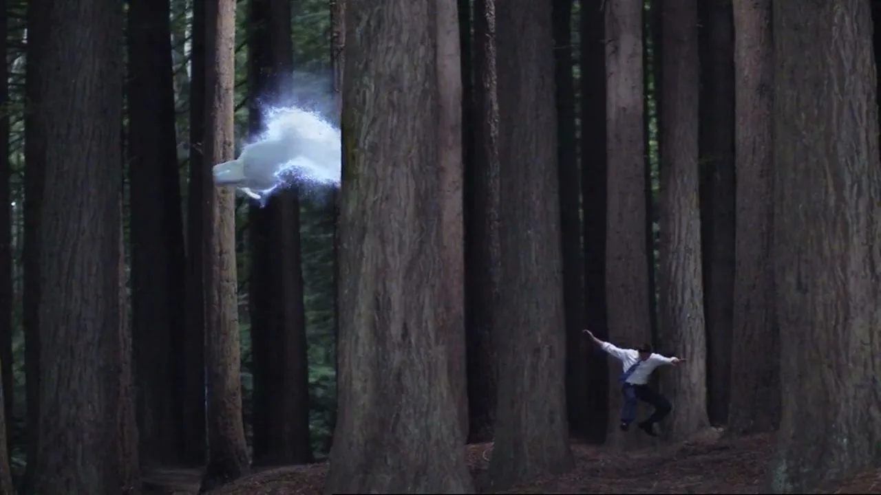 A man in a white shirt and dark pants jumps in a forest of tall trees, while a white, glowing, cloud-like object floats nearby.