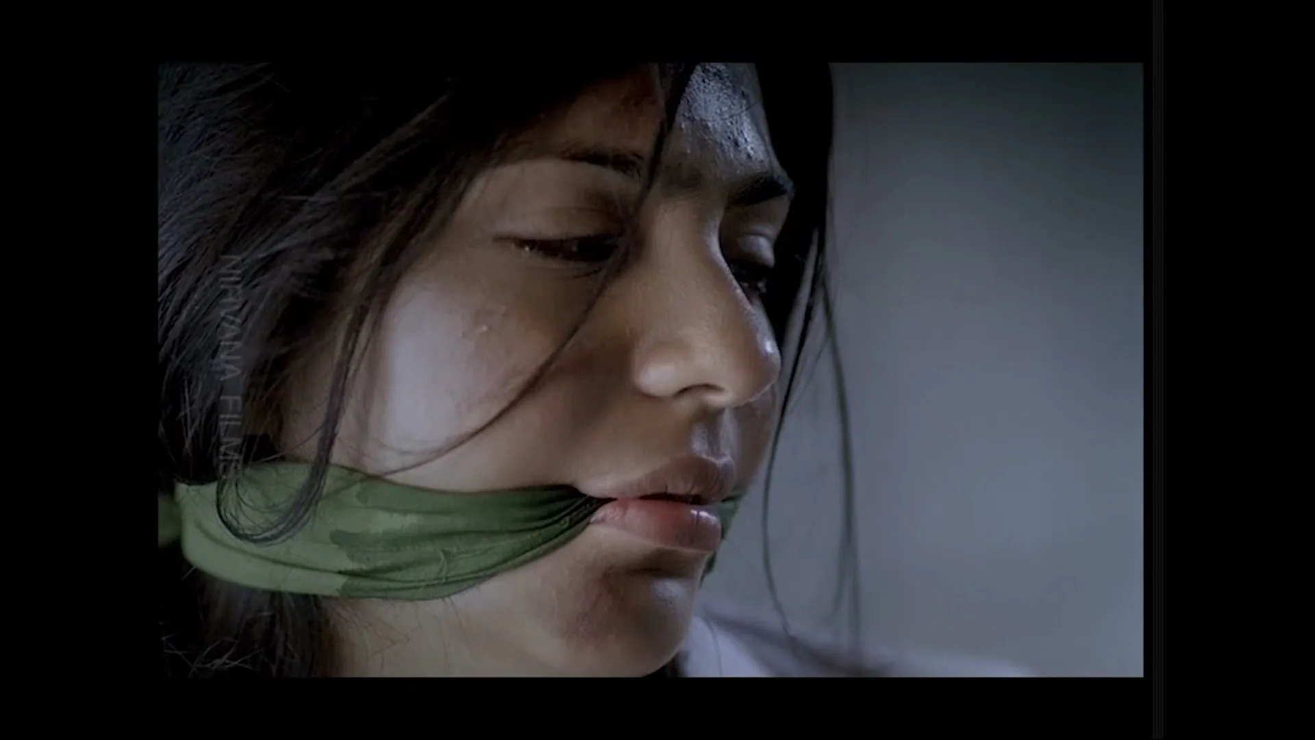 A young woman with dark hair and a green gag over her mouth looks distressed, with visible dirt and sweat on her face, in a dimly lit interior.