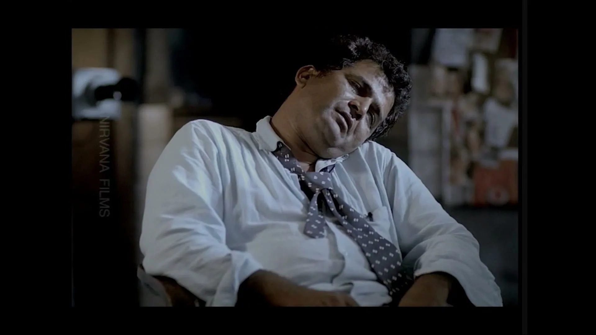 A man in his 40s, wearing a white shirt and a polka-dotted tie, is slumped in a chair, seemingly asleep or unconscious, in a dimly lit room.