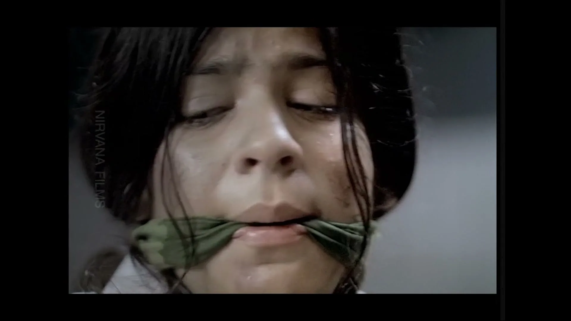 A young woman with dark hair and a green gag over her mouth looks distressed, with tears and dirt on her face, in a dimly lit room.