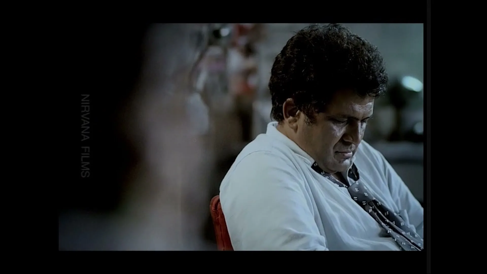 A middle-aged man with curly hair and a white shirt sits slumped, looking down, in a dimly lit interior.