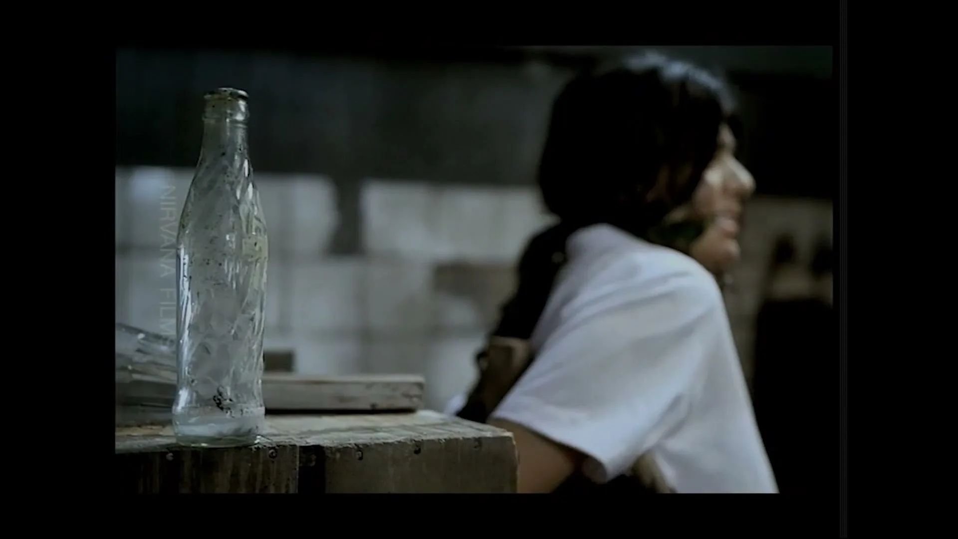 A dirty, empty glass bottle stands on a wooden surface, with a person in a white shirt blurred in the background, looking away.