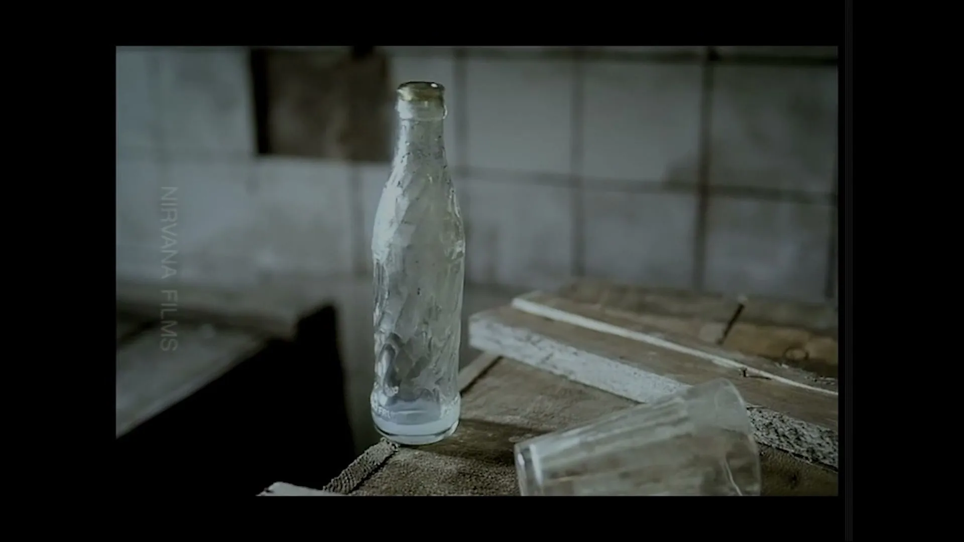 An empty, dirty glass bottle stands upright on a dusty wooden surface next to a fallen glass in a derelict room.