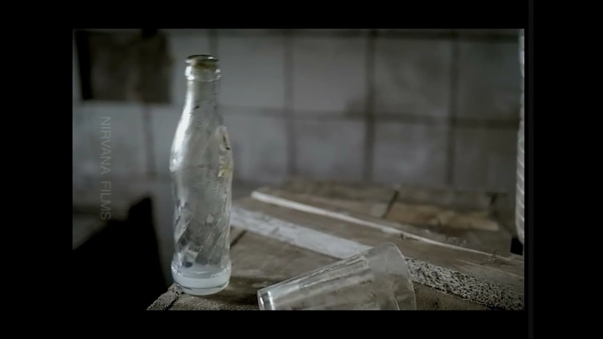 A dirty, empty glass bottle and a broken glass lie on a dusty wooden surface in a dimly lit, abandoned room.