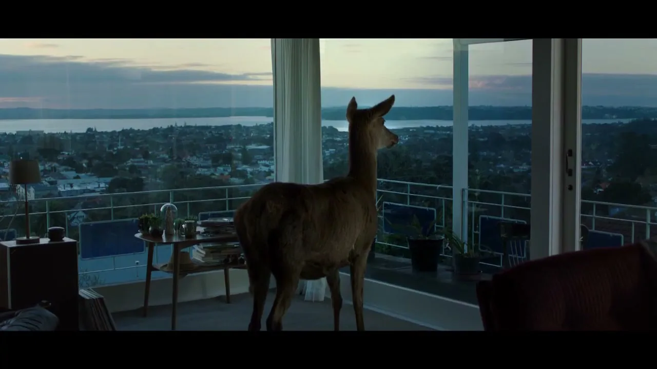 A deer stands inside a modern living room with large windows, looking out at a city and body of water at dusk.