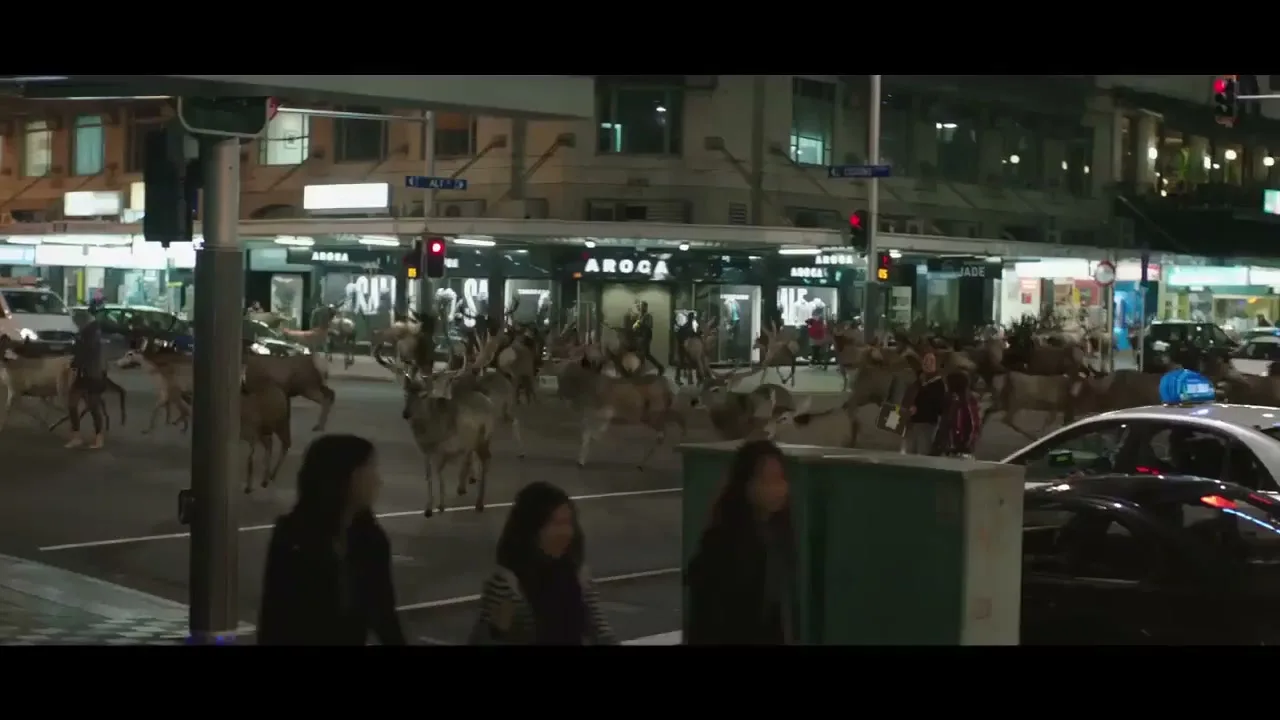 A herd of deer, including several with antlers, runs across a busy city street at night, with cars and pedestrians visible in the background.