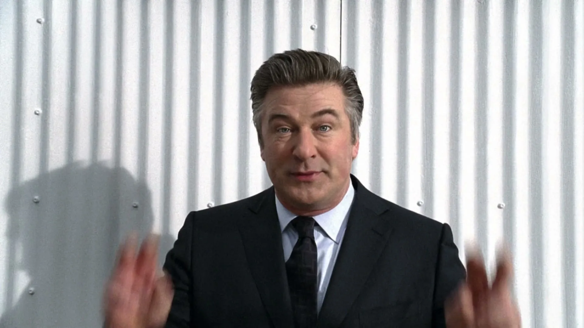 Alec Baldwin, a middle-aged man in a suit, stands in front of a white corrugated wall, talking and gesturing with his hands, then turns and walks away.