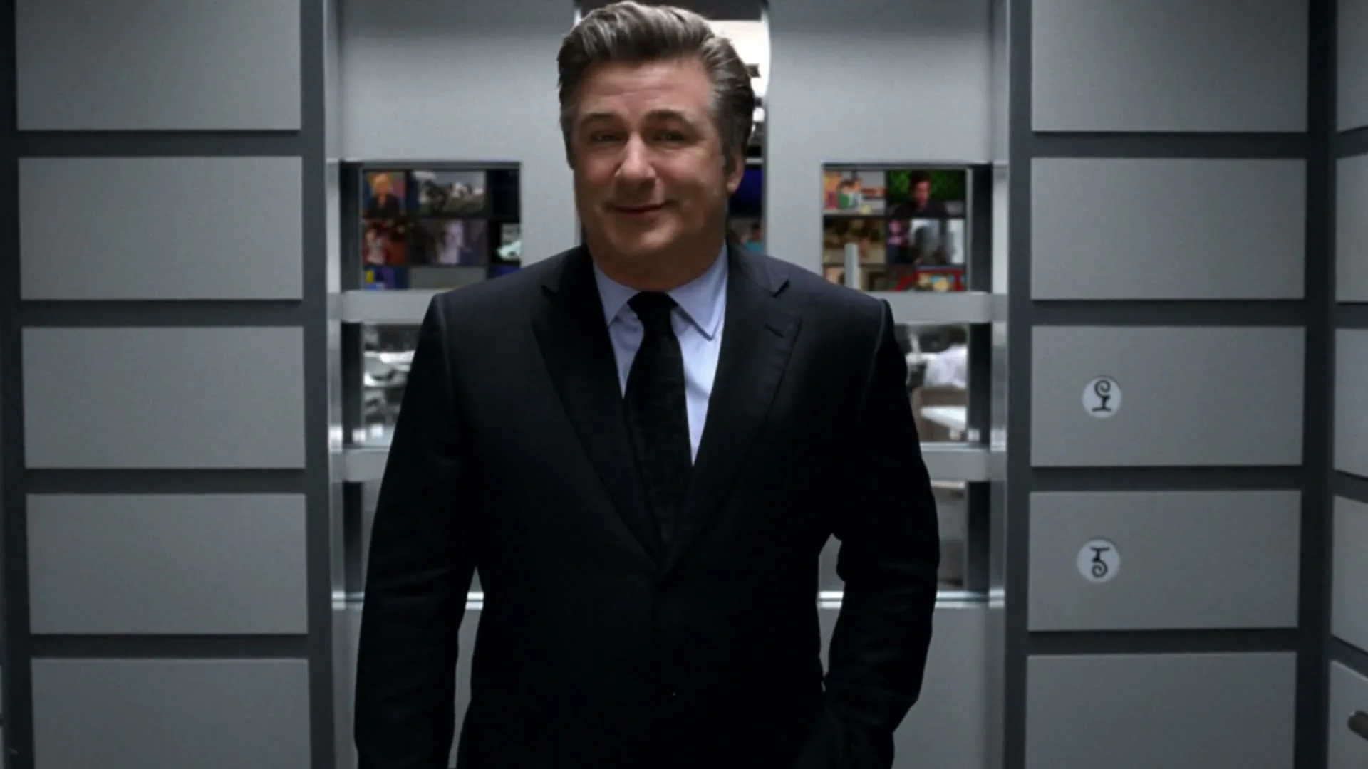 Alec Baldwin, a middle-aged man in a suit, stands in an elevator, then turns and walks out into a control room filled with screens.
