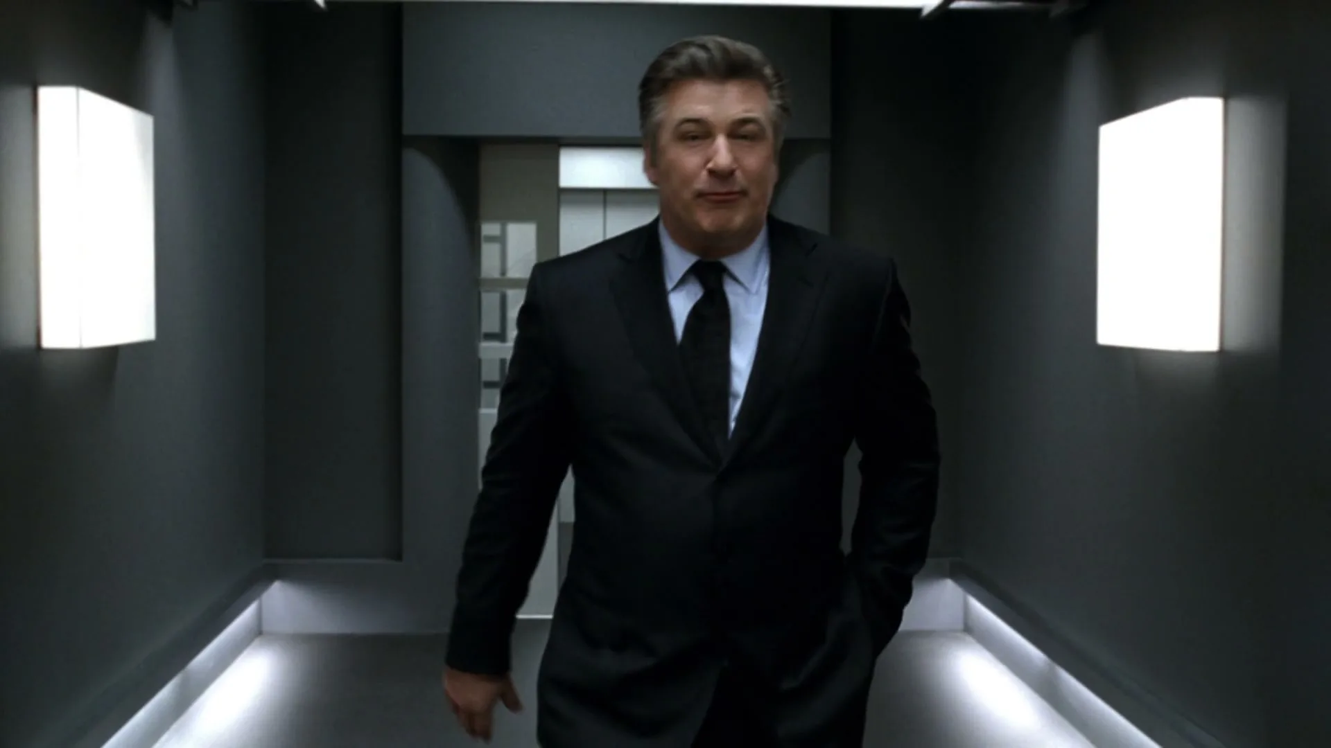 Alec Baldwin, a middle-aged man in a black suit, walks down a futuristic hallway, talking directly to the camera.
