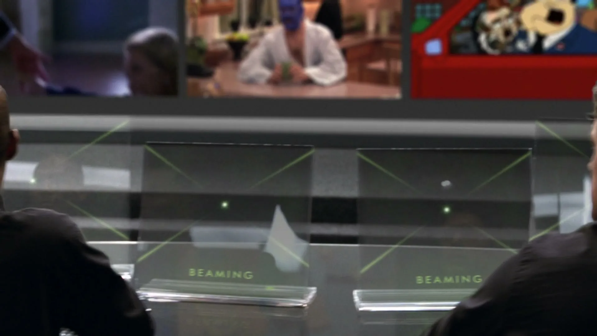 Two men in black shirts sit at a control panel, watching animated characters on multiple screens, with other live-action screens in the background.