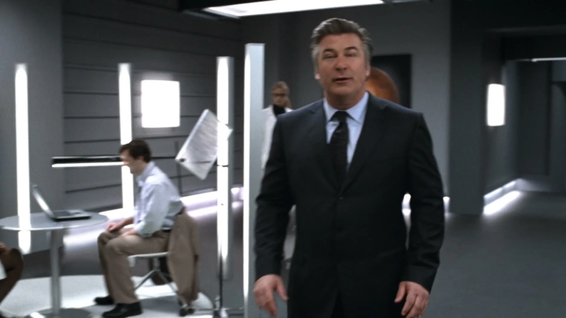 Alec Baldwin, a middle-aged man in a suit, walks towards the camera in a futuristic office, talking to the viewer.