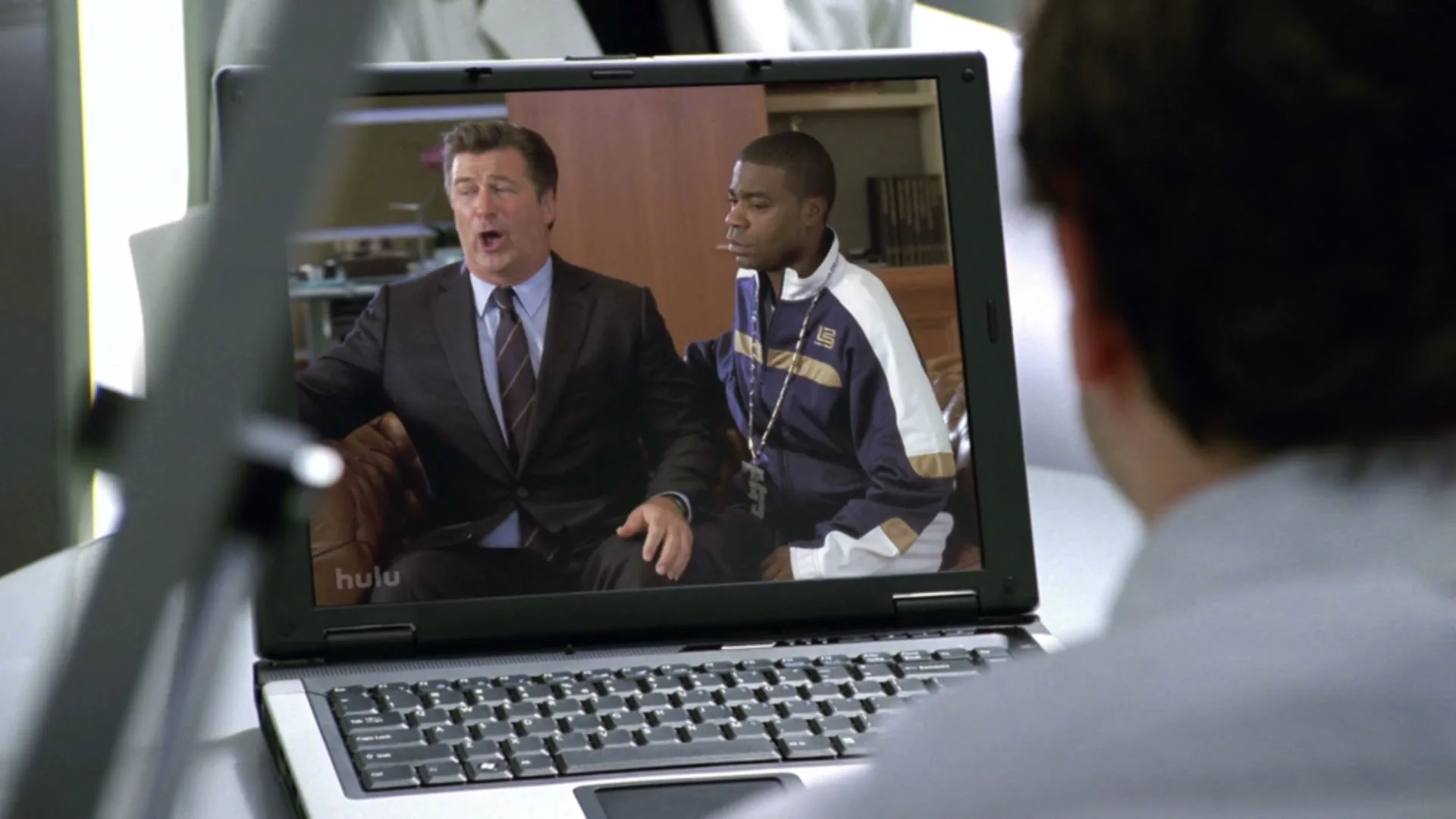 A man watches a video on a laptop of Alec Baldwin and Tracy Morgan sitting on a couch, with Baldwin talking animatedly.