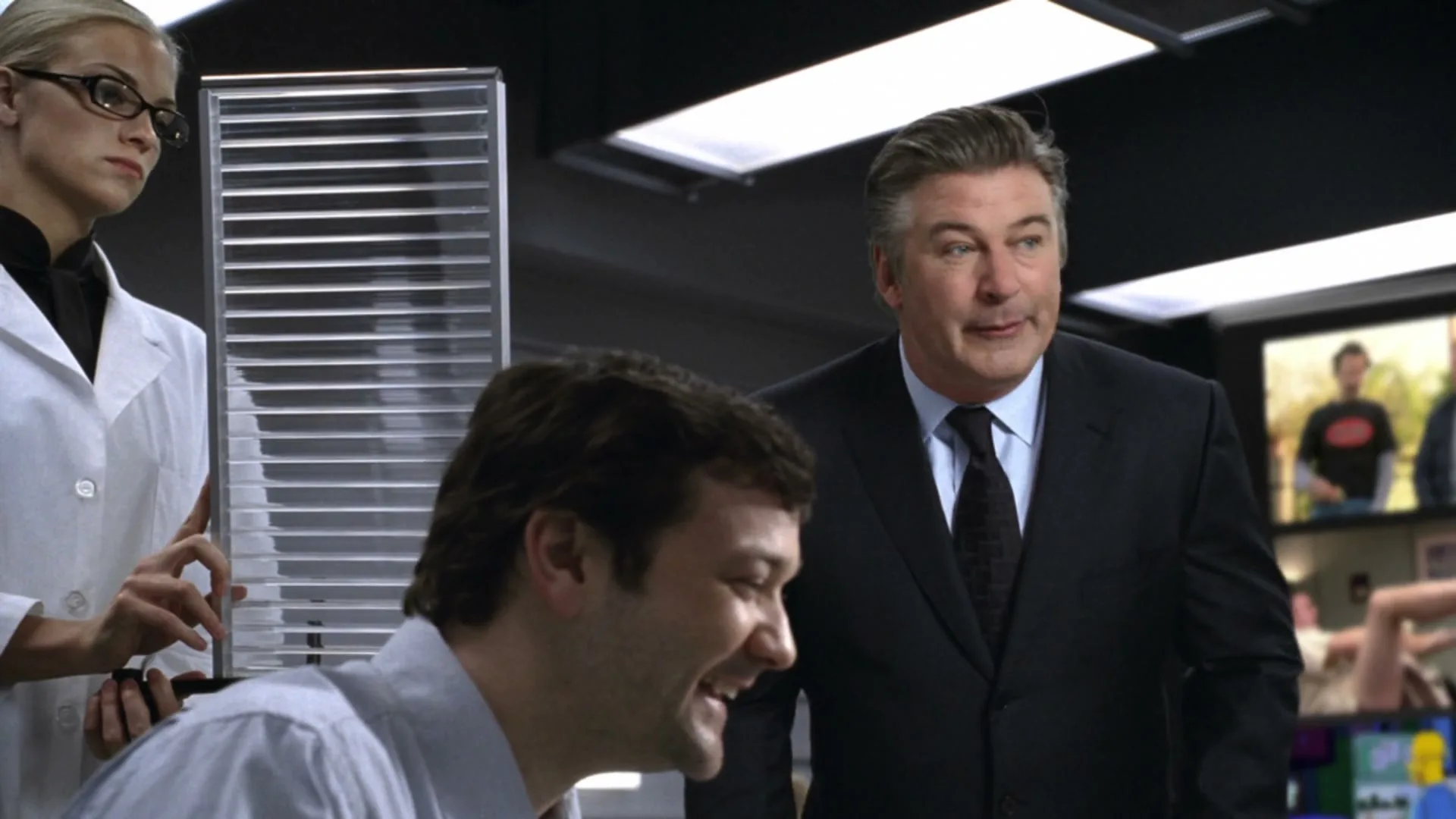 Alec Baldwin, dressed in a suit, stands next to a man in a lab coat who is laughing, while a woman in a lab coat stands nearby in a control room.