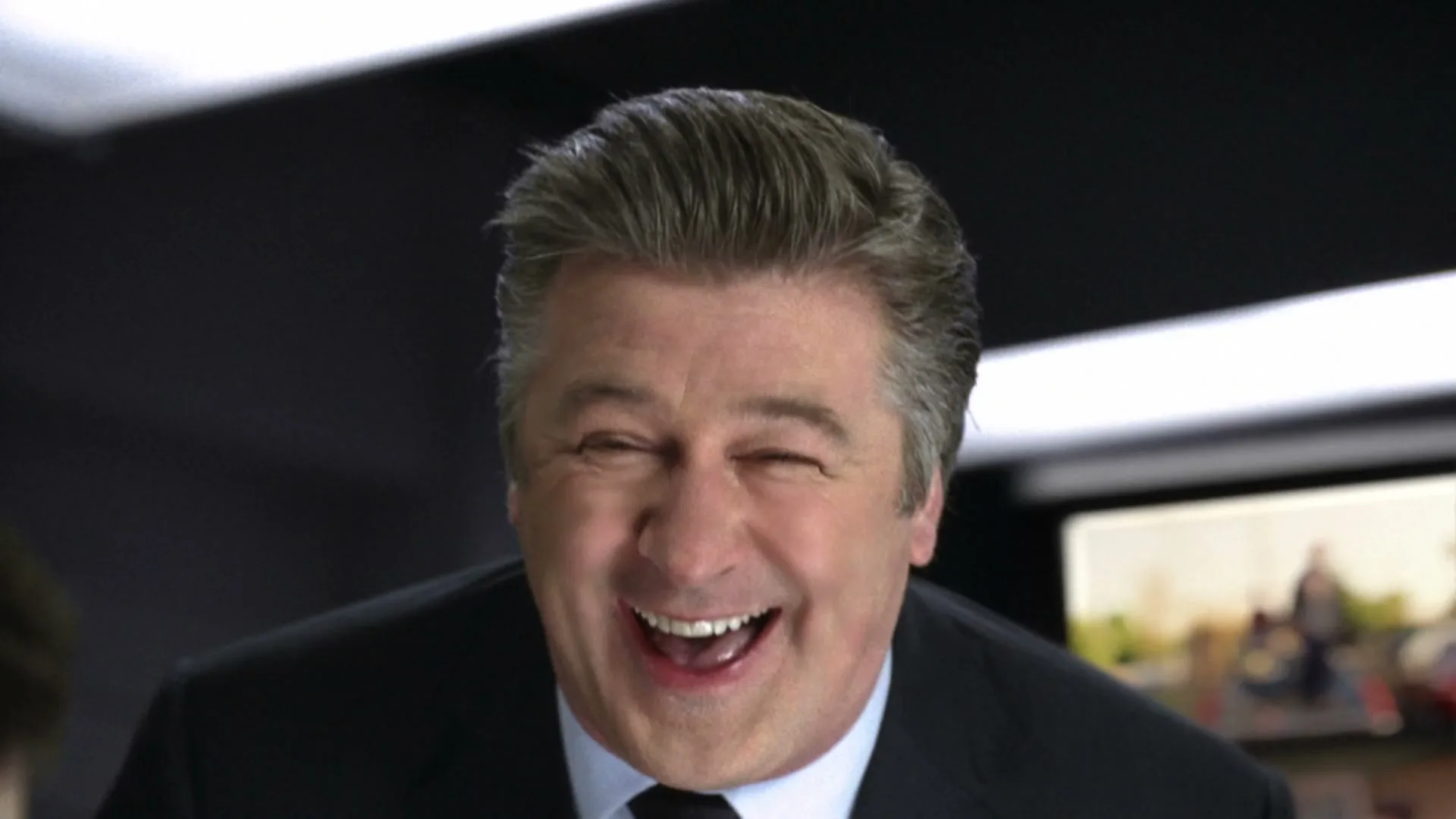 Alec Baldwin, a middle-aged man in a suit, laughs heartily while looking directly at the camera in an office setting.