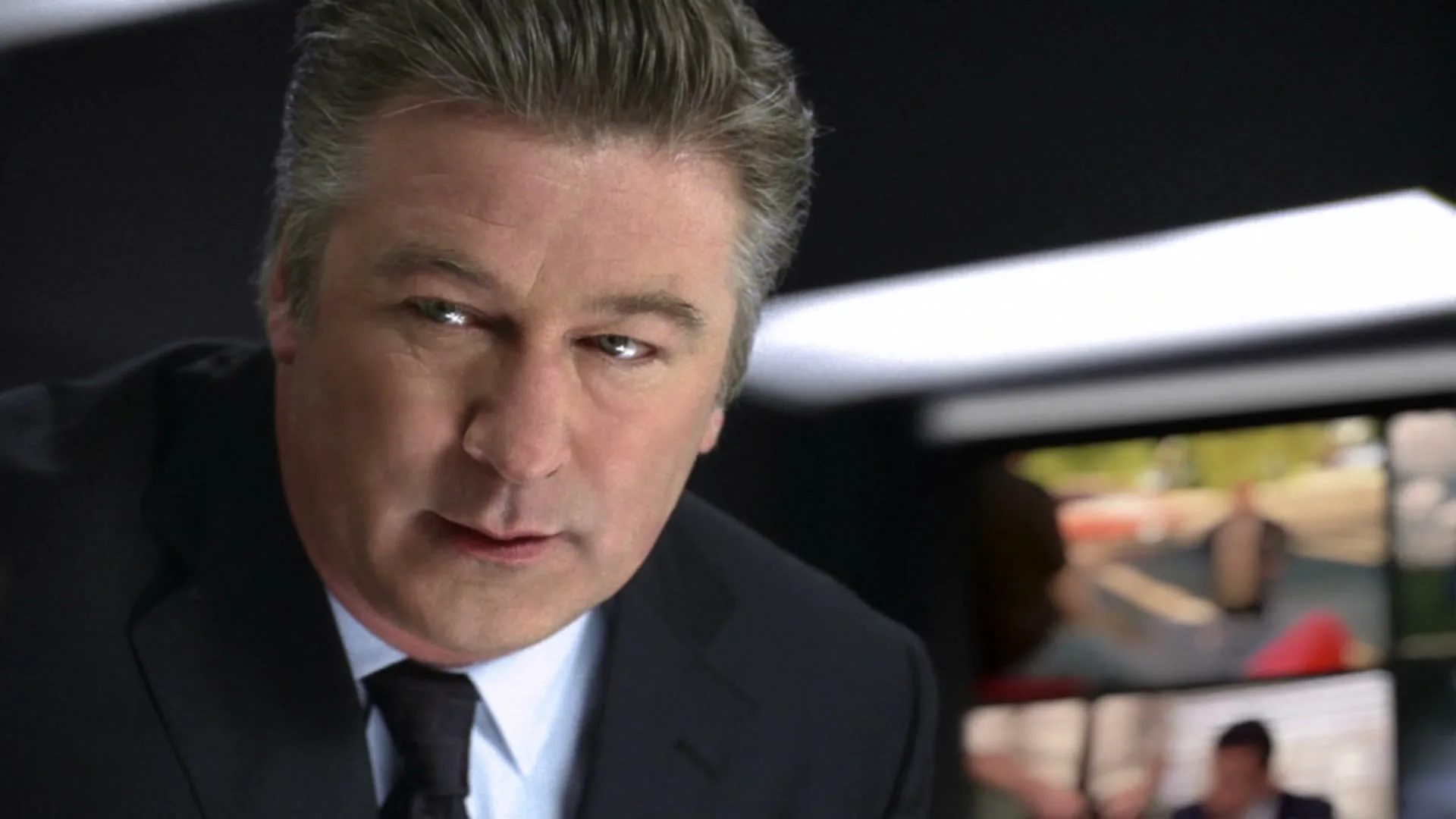 Alec Baldwin, a middle-aged man in a suit, looks intently at the camera in a dark room with multiple screens in the background.