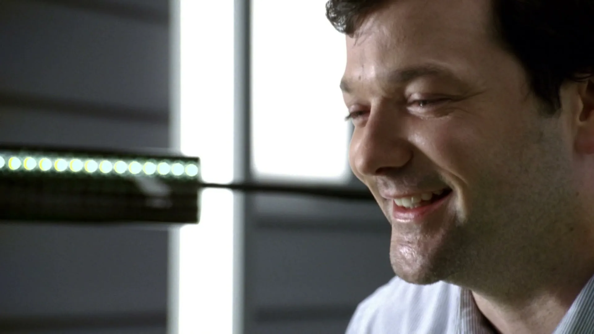 A man with dark hair and a striped shirt smiles, looking off-camera, with a bright LED light fixture in the foreground.
