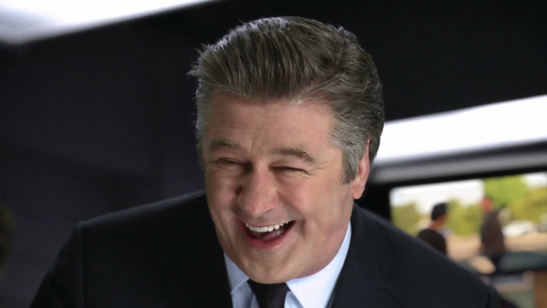 Alec Baldwin, a male in his 50s, wearing a suit, laughs heartily in an interior office setting.