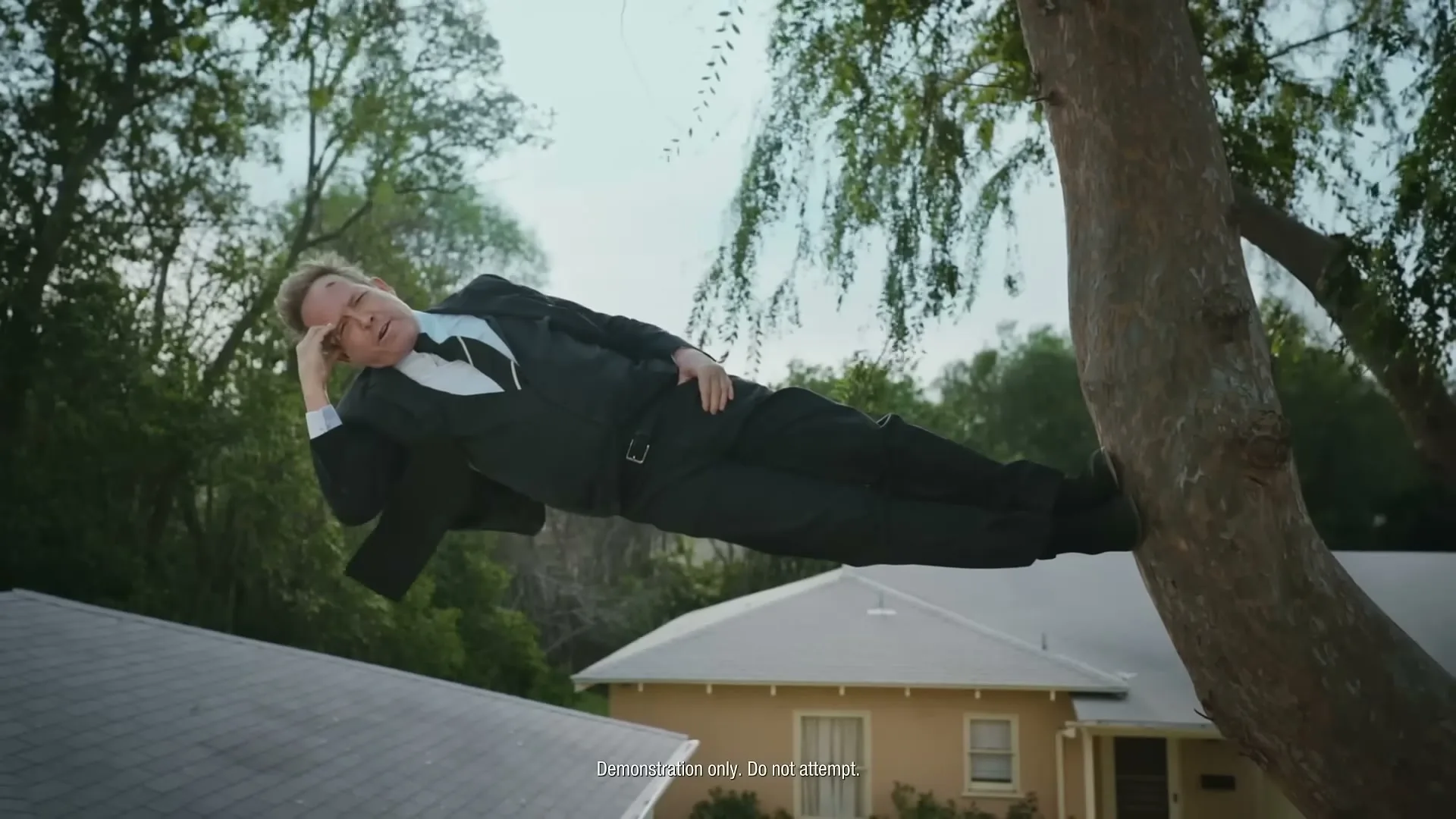 A man in a suit lies horizontally on a tree branch, looking at the camera with a thoughtful expression, above a suburban house.