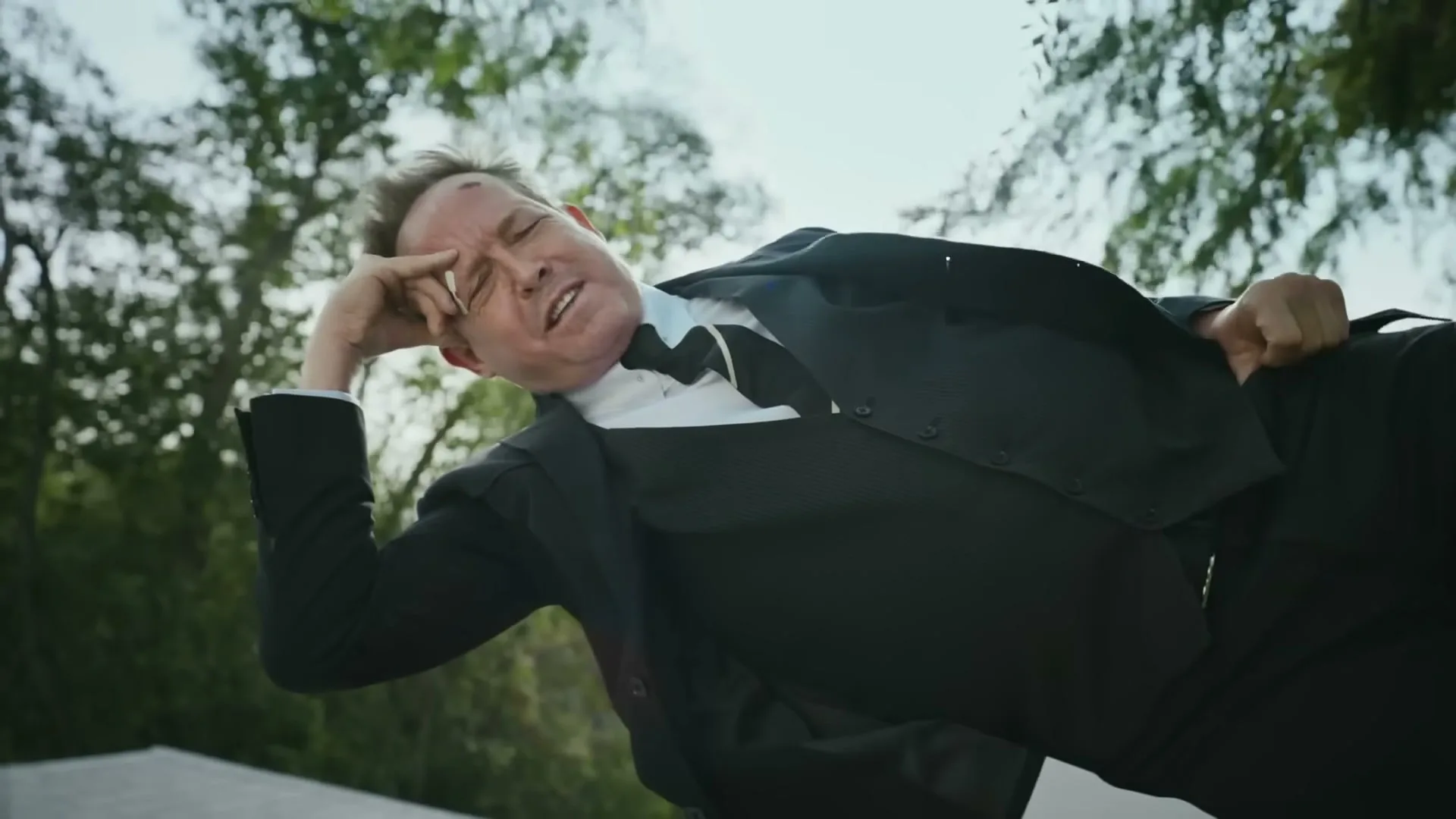 A man in a black suit and white shirt is falling backwards, looking distressed, with trees in the background.