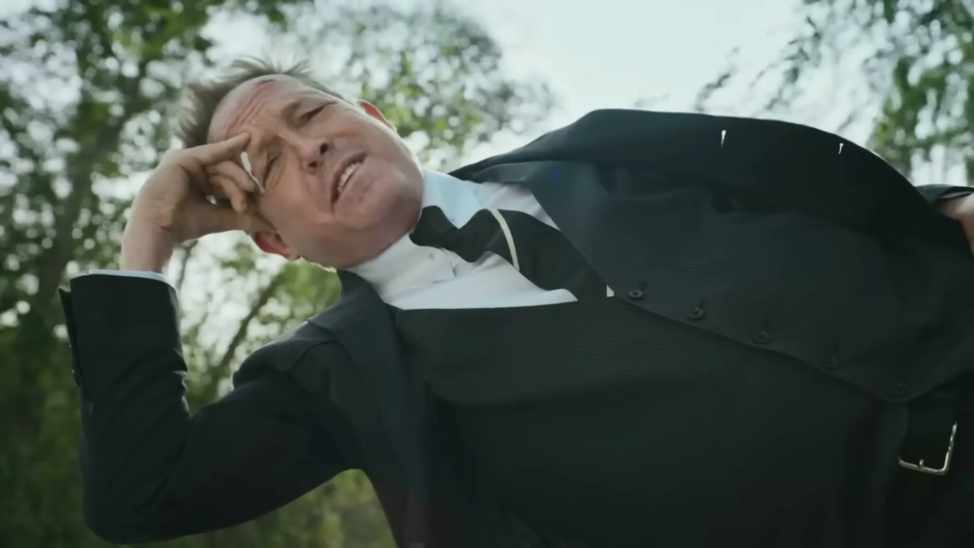 A man in a suit lies on the ground, looking up at the sky with a dazed expression, as the camera pulls back to reveal trees.