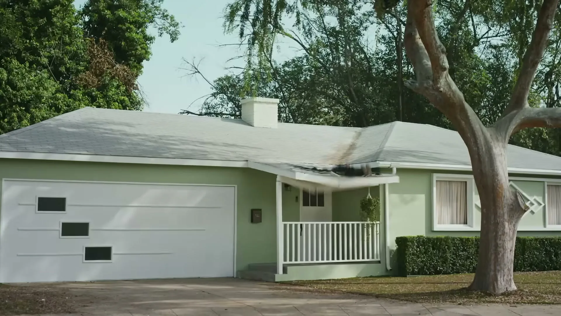 A large tree branch falls from above, crashing onto the porch roof of a light green suburban house, causing significant damage.