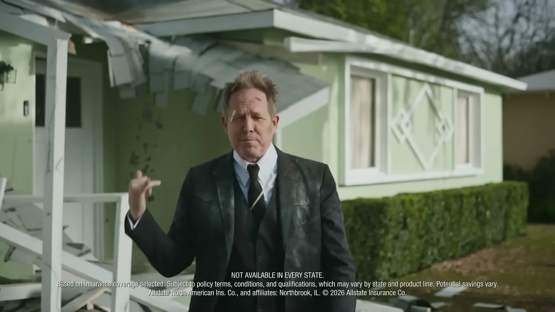 A man in a suit, covered in debris, stands in front of a damaged house, gesturing as he speaks directly to the camera.