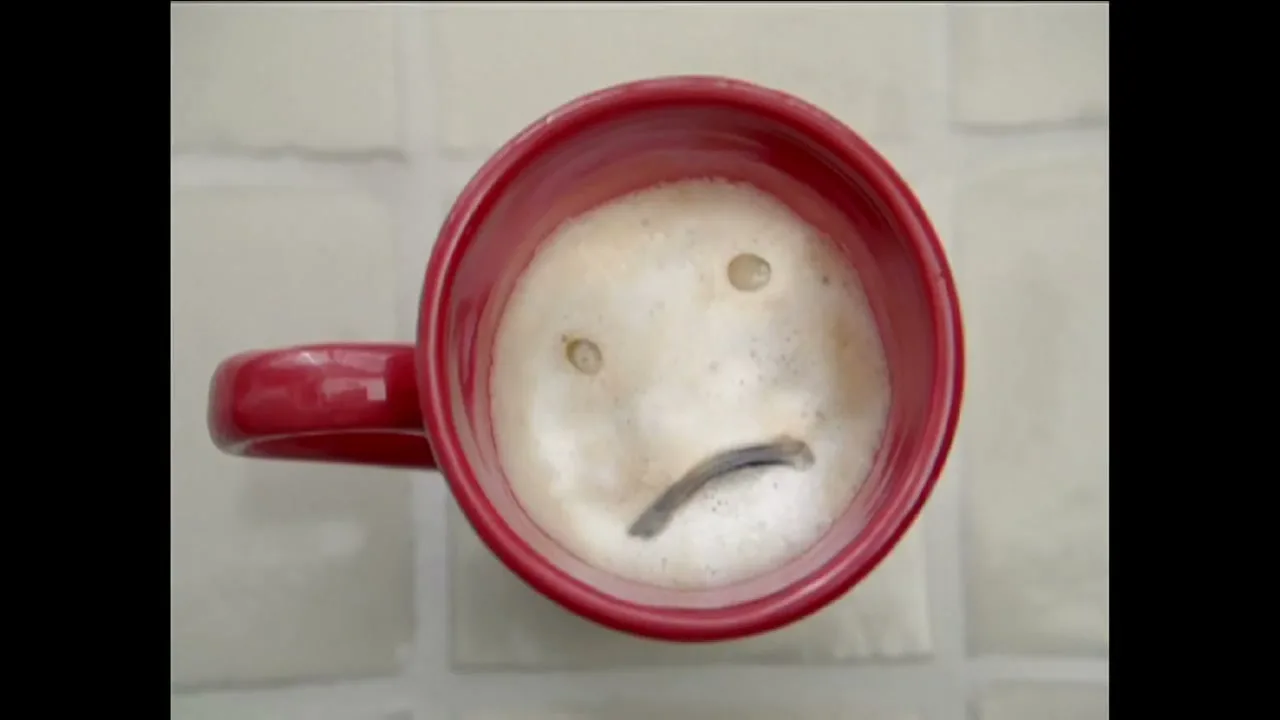 A red mug of coffee with foam on top, forming a sad face with two coffee beans for eyes and a curved piece of chocolate for a mouth, is seen from above on a light-colored surface.