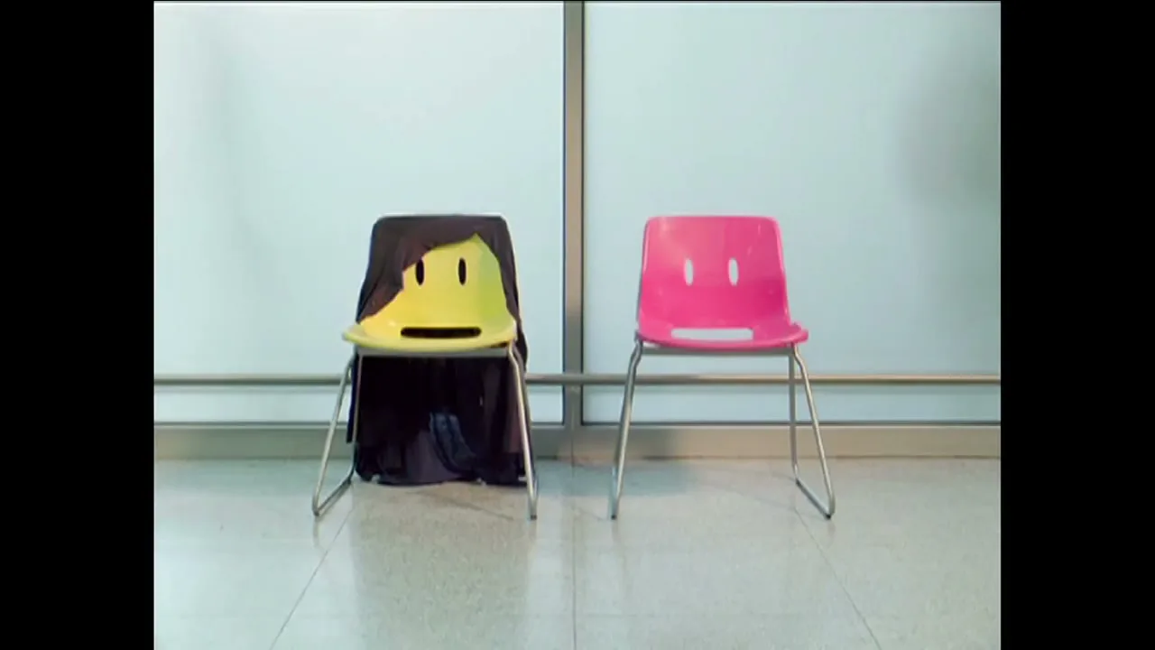 Two empty chairs, one yellow with a black cloth and one pink, sit side-by-side in a brightly lit, minimalist room.