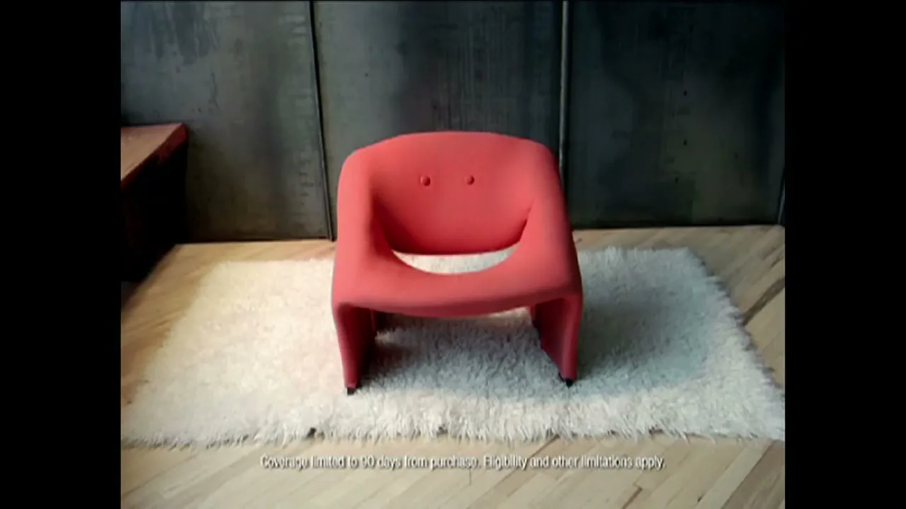 A red chair with a face-like design sits on a white rug in a room with a concrete wall and wooden floor.