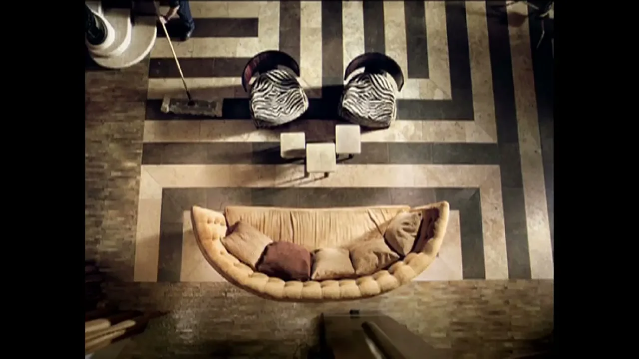 A person mops a patterned floor in a luxurious room with a curved sofa and two chairs, seen from an overhead perspective.