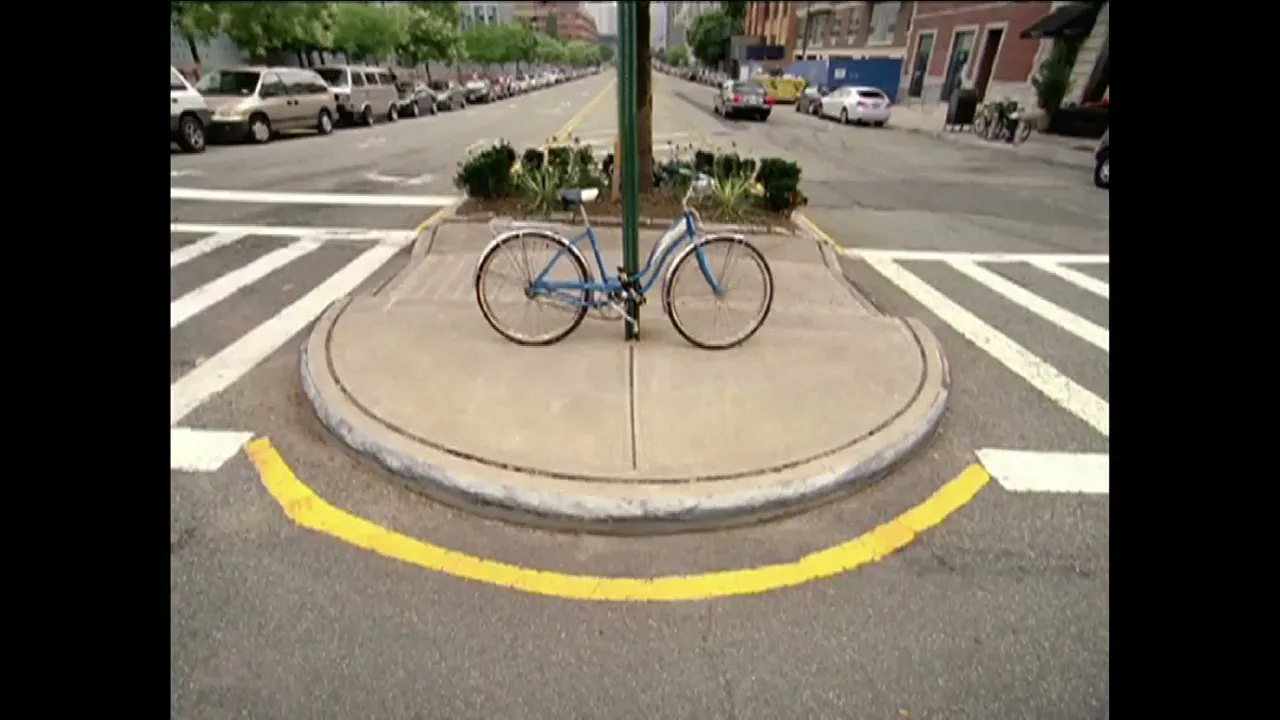 A blue bicycle is parked on a small concrete island in the middle of a city street intersection on a bright day.