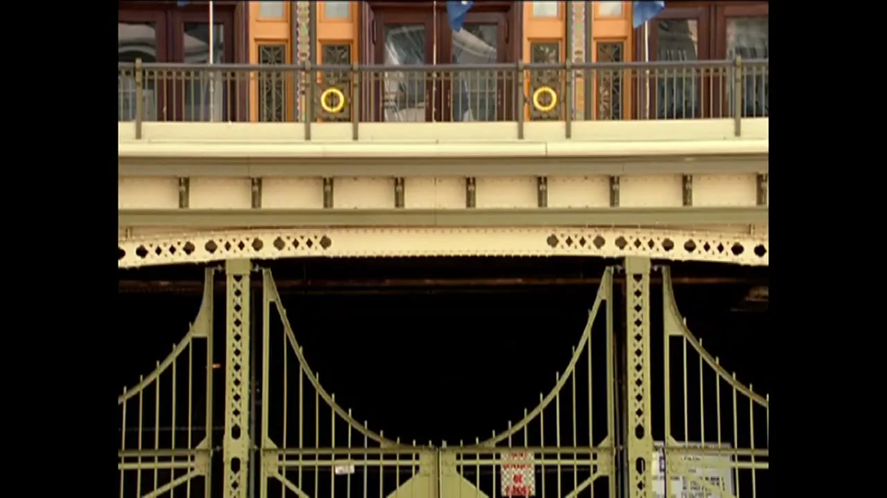 A static shot of a green metal gate and a bridge structure above it, with building windows visible at the top.