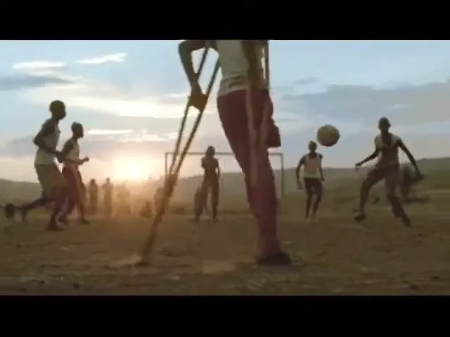 A young boy with crutches plays soccer with other children on a dusty field at sunset, kicking the ball with his foot.