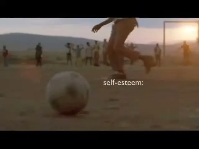 A group of children play soccer on a dusty field at sunset, with one child kicking the ball towards the camera.