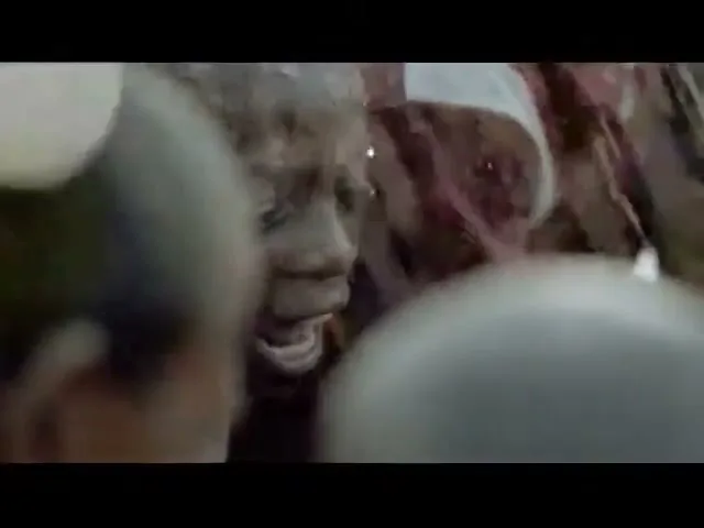 A young boy with dark skin and short hair, wearing a red and white sports jersey, is seen from a low angle, looking up with an excited expression amidst a crowd.